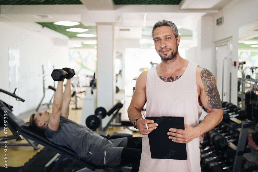 Muscular handsome trainer writing a fitness plan on clipboard for working out near his trainee in the fitness