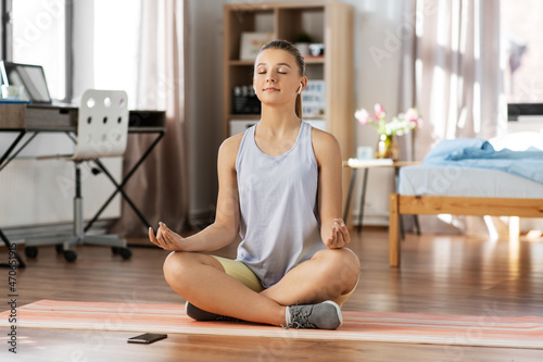 Fototapeta Naklejka Na Ścianę i Meble -  sport, fitness and healthy lifestyle concept - happy teenage girl with smartphone and earphones sitting on yoga mat and meditating at home