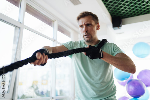 Caucasian blonde man training with rope in functional training fitness gym with big windows and a lot of daylight.