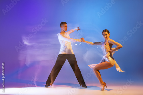 Two young graceful dancers, flexible man and woman dancing ballroom dance isolated on gradient blue purple background in neon mixed light
