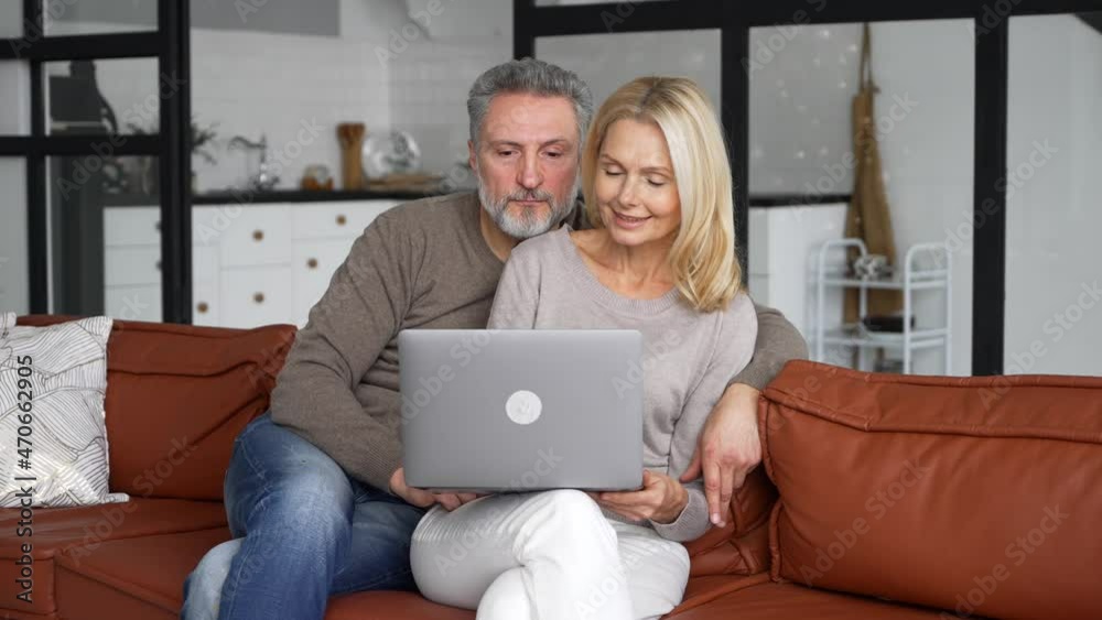 Middle-aged couple in love using laptop sitting on the couch at home. Cheerful senior couple watching movie together, smiling and hugging at cozy living room, web surfing and spends time online Middle-aged couple in love using laptop sitting on the couch at home. Cheerful senior couple watching movie together, smiling and hugging at cozy living room, web surfing and spends time online