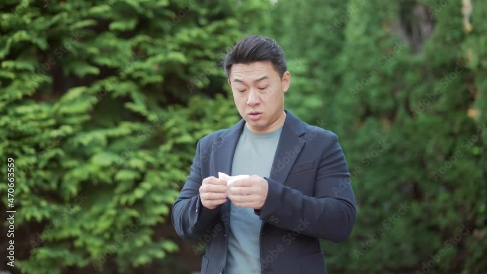 portrait of a sick young asian man who sneezes and coughs uses a handkerchief on the street outdoors. Business Male in a suit suffers from season allergies unhealthy flu while standing outside portrait of a sick young asian man who sneezes and coughs uses a handkerchief on the street outdoors. Business Male in a suit suffers from season allergies unhealthy flu while standing outside