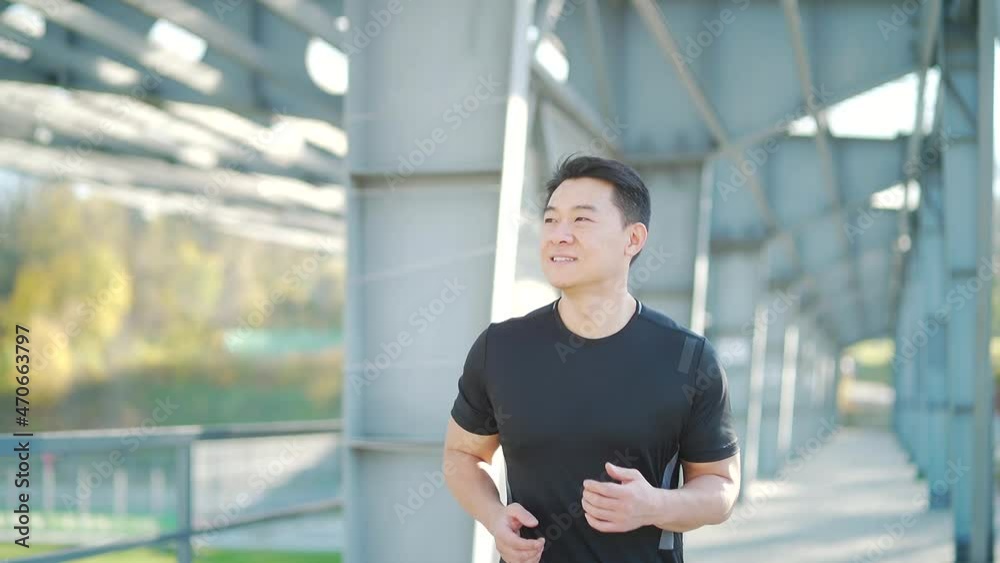 young asian runner running on modern urban stadium background. Morning jog sports Athletic man in black sportswear. jogging down the city street. Outdoors bridge. slow motion. Healthy Lifestyle young asian runner running on modern urban stadium background. Morning jog sports Athletic man in black sportswear. jogging down the city street. Outdoors bridge. slow motion. Healthy Lifestyle