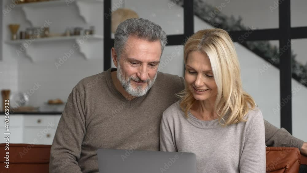 Senior couple is using laptop for watching sports match or online lottery together sitting on comfortable sofa in living room. Cheerful middle-aged spouses raising hands up in victory gesture, triumph Senior couple is using laptop for watching sports match or online lottery together sitting on comfortable sofa in living room. Cheerful middle-aged spouses raising hands up in victory gesture, triumph