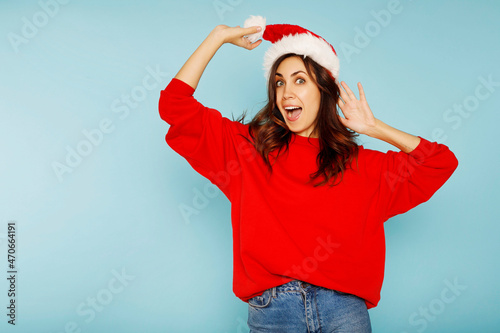 Young american woman wearing and holding christmas red Santa Claus hat with hand near face, shocked and surprised. Christmas and New Year news concept.