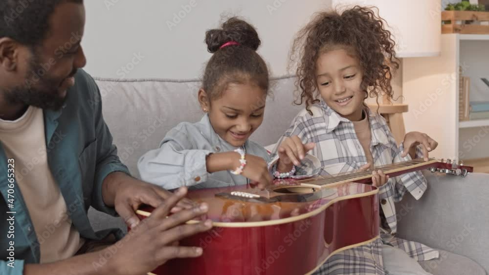 Medium slowmo shot of two funny little sisters and their African-American father sitting on sofa at cozy home having fun together playing guitar Medium slowmo shot of two funny little sisters and their African-American father sitting on sofa at cozy home having fun together playing guitar