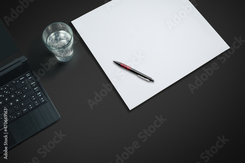 laptop on a dark desk with paper and pen and  a glas of sparkling water
