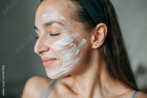 A brunette woman 30 years old with a beam on her head in underwear stands in the bathroom and enjoys with her eyes closed with a cream on her face
