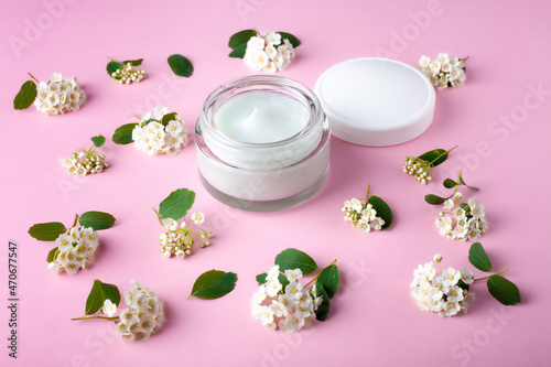 Glass jar and white cap with body cream on pink background with small white flowers. Transparent jar with face cream isolated. Cosmetic product for skin care.