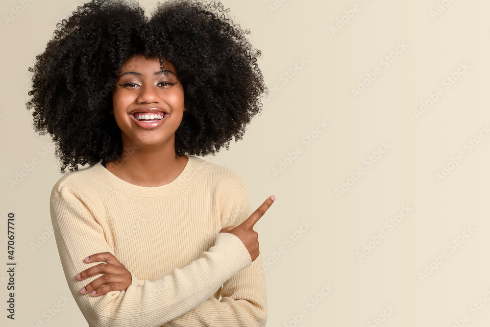 Smiling dark-skinned young woman points with her finger to a space on the right side of the image, isolated on beige background. Smiling dark-skinned young woman points with her finger to a space on the right side of the image, isolated on beige background.