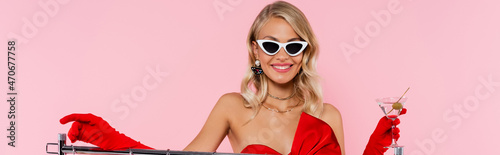 cheerful woman in stylish sunglasses and red gloves holding cocktail while smiling isolated on pink, banner