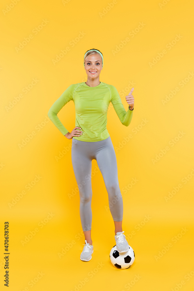 smiling sportswoman with hand on hip showing thumb up while stepping on soccer ball on yellow smiling sportswoman with hand on hip showing thumb up while stepping on soccer ball on yellow