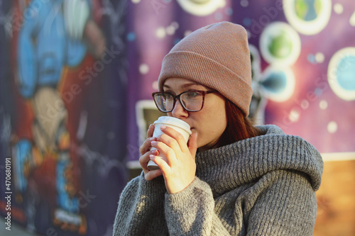 A young girl in glasses with a disposable paper cup drinks fresh coffee in the park against a background of purple graffiti © Сергей Стельченко