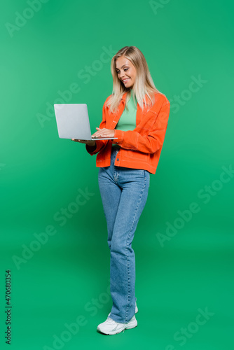 full length view of smiling woman in orange jacket and jeans using laptop on green