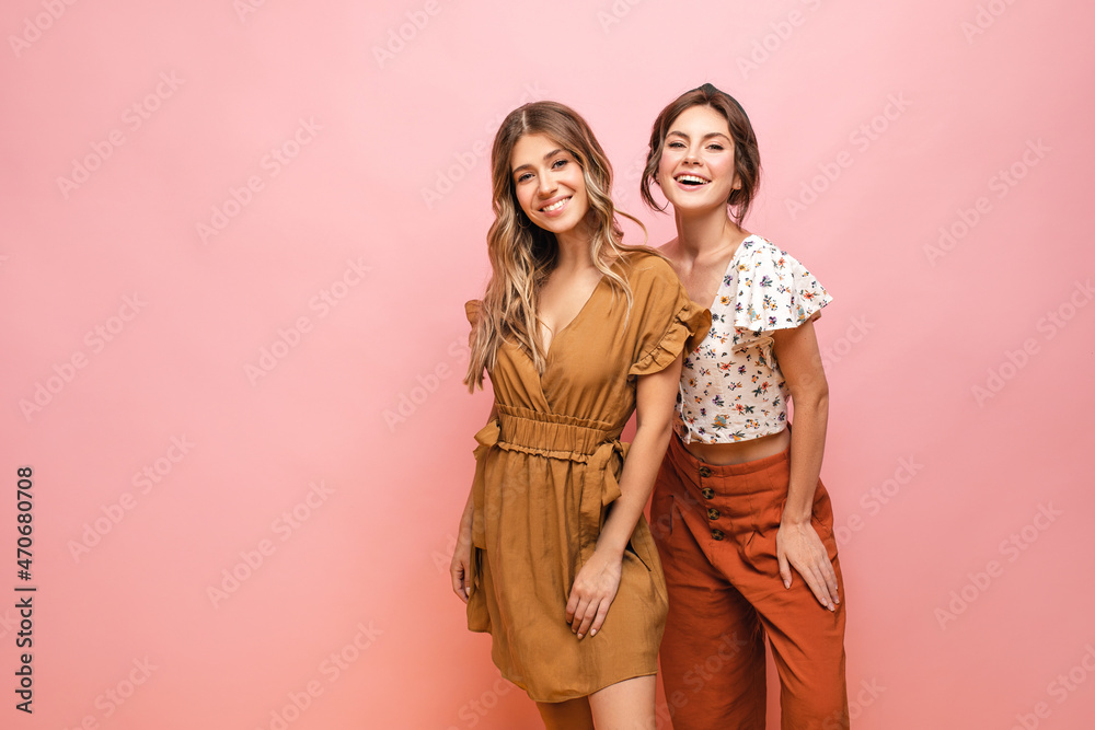 Two pretty young caucasian ladies standing side by side smiling at camera against pink background. Blonde with hair down wearing dress. Brown-haired woman with gathered hairstyle in blouse, trousers. Two pretty young caucasian ladies standing side by side smiling at camera against pink background. Blonde with hair down wearing dress. Brown-haired woman with gathered hairstyle in blouse, trousers.