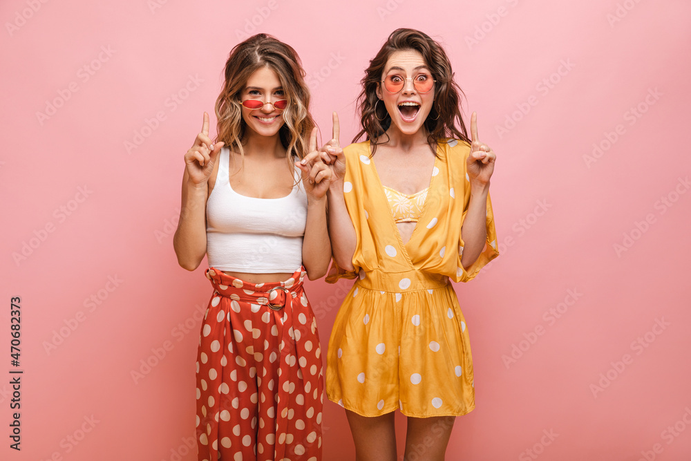 Cute caucasian young ladies pointing up with index fingers over isolated pink background. Smiling teenagers in translucent glasses are dressed in light summer clothes. Advertising concept Cute caucasian young ladies pointing up with index fingers over isolated pink background. Smiling teenagers in translucent glasses are dressed in light summer clothes. Advertising concept
