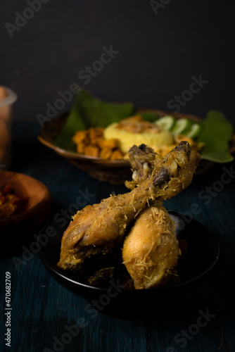 Fried chicken with serundeng (fried coconut) in dark mood theme. Behind: turmeric rice and spicy sauve osr known as sambal and crackers