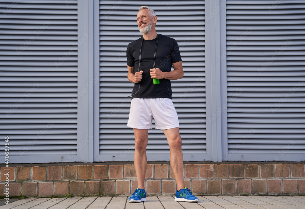 Cheerful middle aged athletic man in sportswear smiling aside while holding skipping rope, standing outdoors over grey background