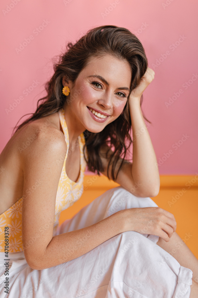 Lovely caucasian young girl with pleasant appearance sits sideways on floor in room with pink wall. On colored background, cute smiling brown-haired woman in summer light clothes. Lovely caucasian young girl with pleasant appearance sits sideways on floor in room with pink wall. On colored background, cute smiling brown-haired woman in summer light clothes.