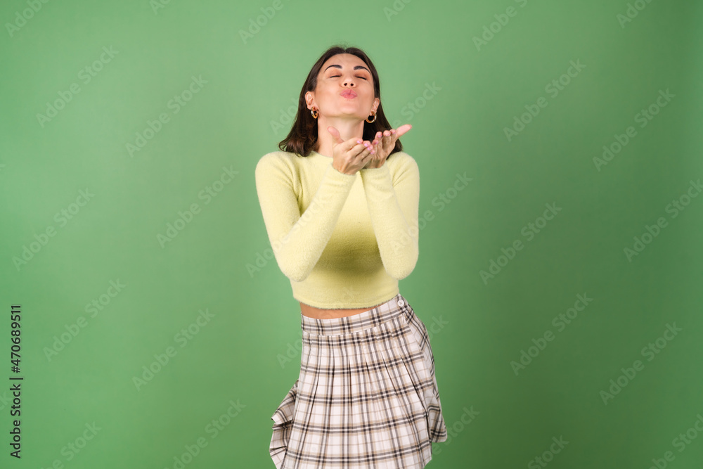 Young woman on a green background in a yellow sweater and a plaid skirt smiles cutely, moves in a good elated mood Young woman on a green background in a yellow sweater and a plaid skirt smiles cutely, moves in a good elated mood