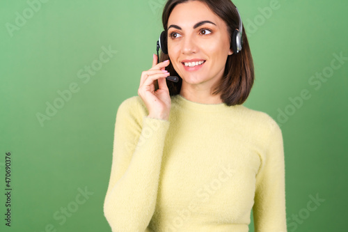 Young woman on a green background in a yellow sweater manager, support service worker, salesperson, with pleasure accepts calls from customers