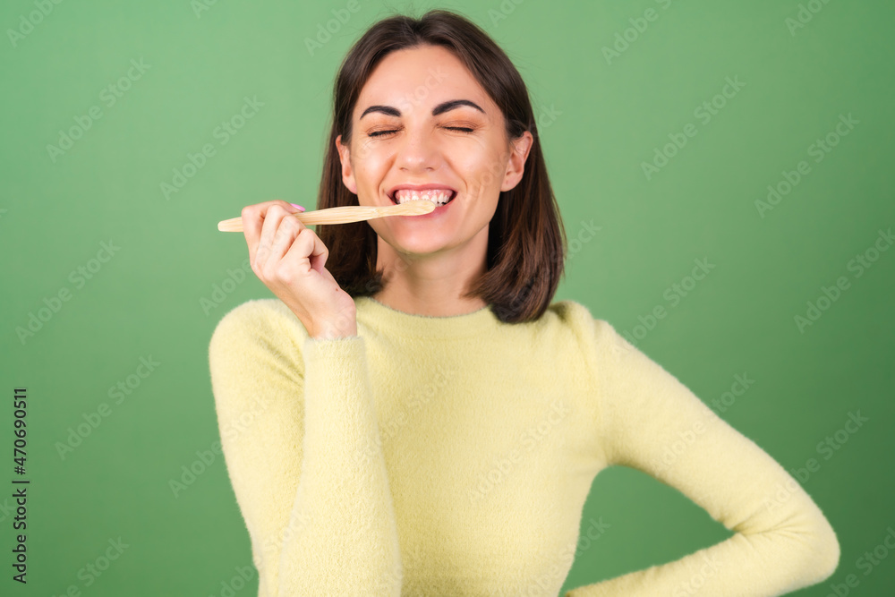 Young woman on a green background in a yellow sweater holds an ecological wooden toothbrush, taking care of the nature and a tooth Young woman on a green background in a yellow sweater holds an ecological wooden toothbrush, taking care of the nature and a tooth