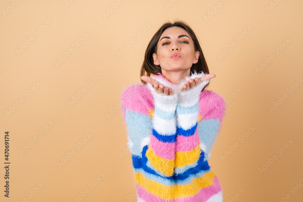 Young woman on a beige background in a bright multi-colored cozy knitted sweater cute sends an air kiss Young woman on a beige background in a bright multi-colored cozy knitted sweater cute sends an air kiss
