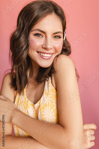 Close-up of caucasian young girl with snow-white smile on her face stands on pink background hugging herself. Beauty with wavy brown hair, light makeup and yellow T-shirt.