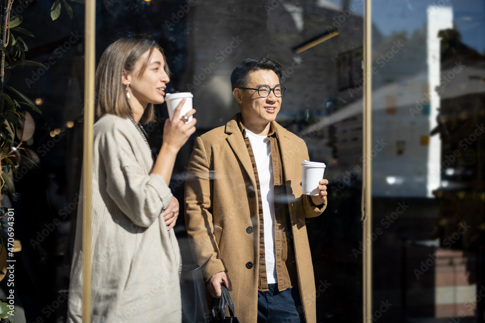 Asian businessman and caucasian businesswoman going, talking and drinking coffee inside unknown building. Concept of remote and freelance work. Idea of teamwork and business cooperation. Sunny day Asian businessman and caucasian businesswoman going, talking and drinking coffee inside unknown building. Concept of remote and freelance work. Idea of teamwork and business cooperation. Sunny day