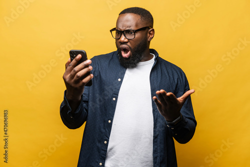 Shocked surprised African-American man staring at smartphone with wide open mouth isolated on yellow background. Black guy feels negative emotions, amazed facial expression