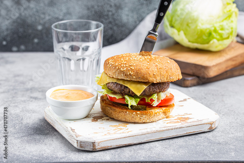 Delicious burger pierced with knife with meat beef patty, cheese Cheddar, salad and fresh tomato served with sauce on white wooden cutting board and cola in tall glass