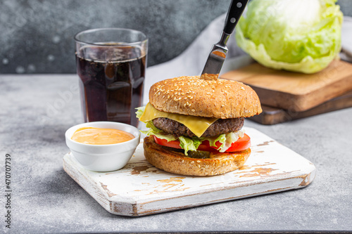 Delicious burger pierced with knife with meat beef patty, cheese Cheddar, salad and fresh tomato served with sauce on white wooden cutting board and empty tall glass