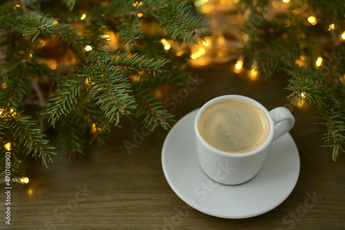 Christmas or winter coffee. Coffee cup, Christmas tree and Christmas lights, golden bokeh, blurred background. Black coffee atmosphere of winter festive and hot aroma drink at a wintertime, new year