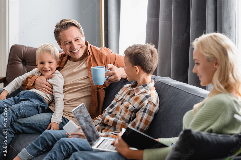 happy man with cup of tea sitting on sofa with sons while blurred wife working on laptop