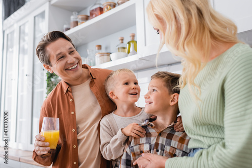 cheerful man with orange juice looking at cheerful sons and wife in kitchen