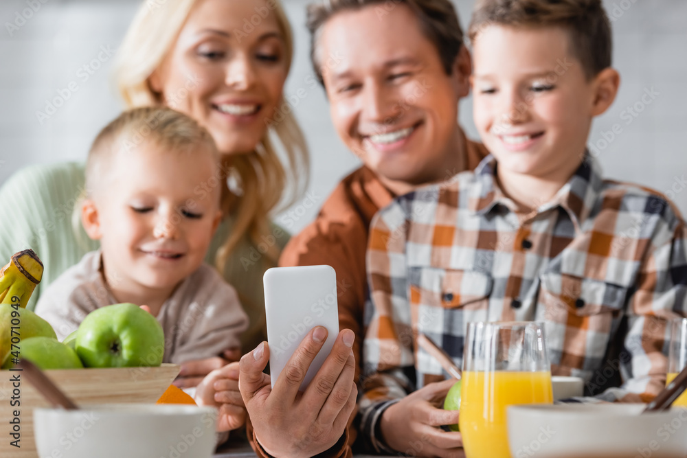 man taking selfie on smartphone near blurred happy family during breakfast man taking selfie on smartphone near blurred happy family during breakfast