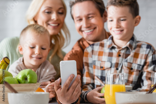 man taking selfie on smartphone near blurred happy family during breakfast