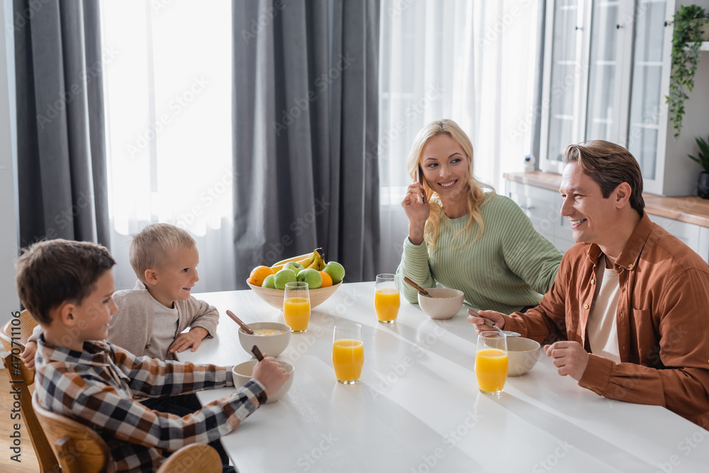 smiling woman talking on smartphone during breakfast with husband and sons