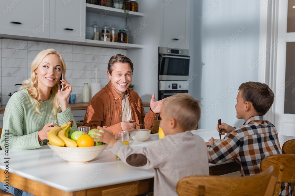 cheerful man talking to sons during breakfast while wife calling on mobile phone