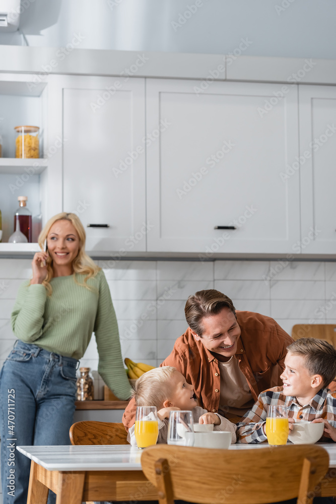 blurred and happy woman talking on mobile phone near husband with sons having breakfast in kitchen