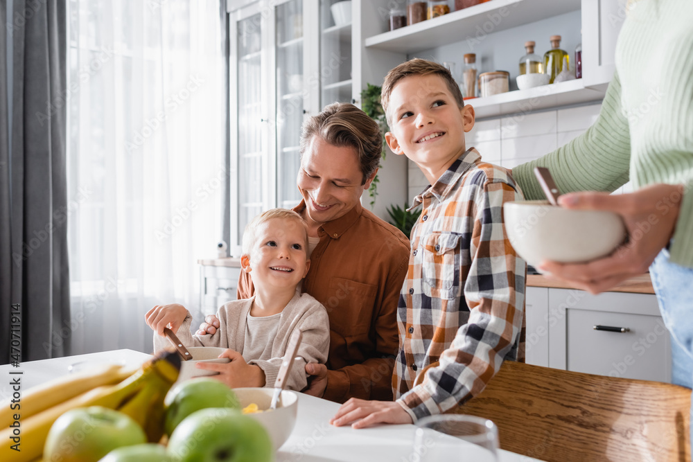 father with boys having breakfast near blurred fruits and mother with bowl father with boys having breakfast near blurred fruits and mother with bowl