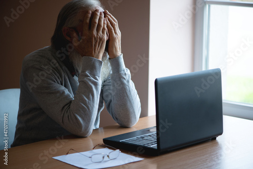 A tired, broken, resigned, preoccupied elderly man sits in front of a laptop, computer and covers his face with his hand. The sad senior checks his account balance.