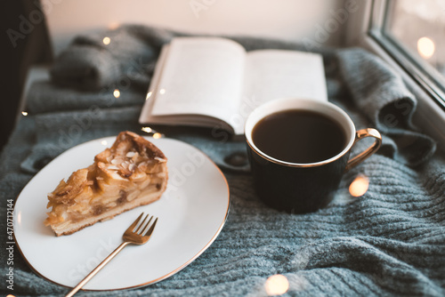 Tasty apple pie on white plate with cup of black tea and open paper book on knit woolen cloth fabric closeup over Christmas glow lights. Winter holiday season. Cozy home atmosphere. Coffee break.