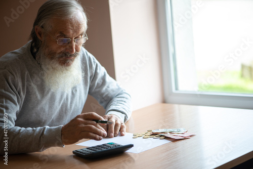 Old bearded senior man with calculator and bills counting euro money at home.