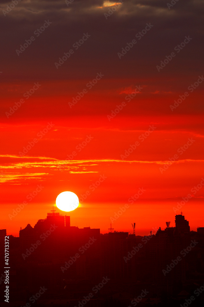 A bright red sun disk rises in the early morning over the silhouettes of city houses. Vertical image.