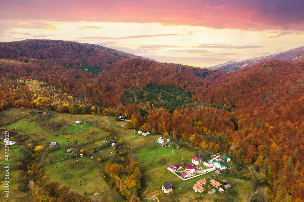Aerial view of small shepherd houses on wide meadow between autumn forest in Ukrainian Carpathian mountains at sunset