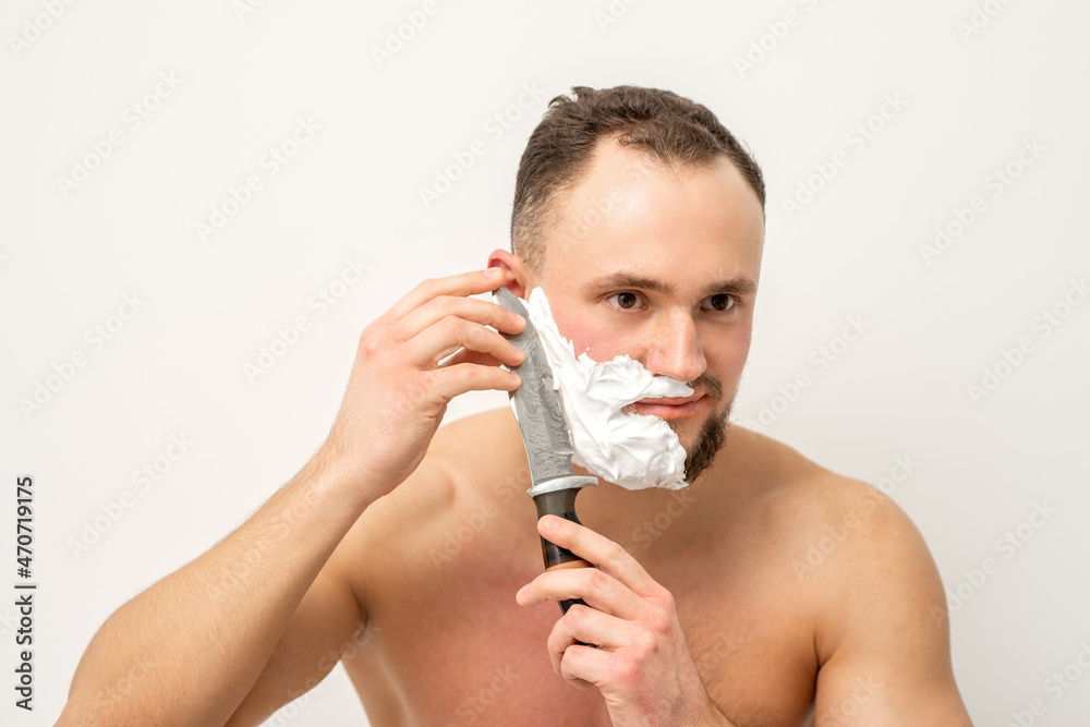 Young caucasian man shaving beard with a big knife on white background.