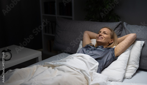 Pleasant mature woman in pajamas lying on comfy bed with soft linen and enjoying relaxation time. Caucasian blonde smiling and keeping hands behind head.