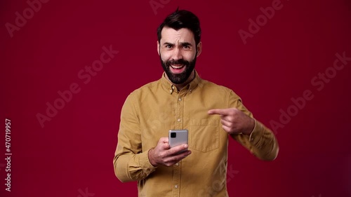 the guy looks at the phone and shows a like at him, surprised, he is bearded, black-haired, dressed in a brown shirt, the background is red