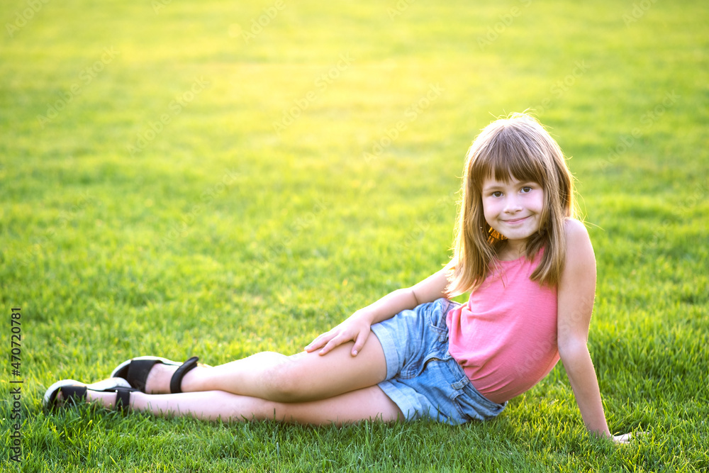 Young happy child girl resting while sitting on green grass lawn on warm summer day